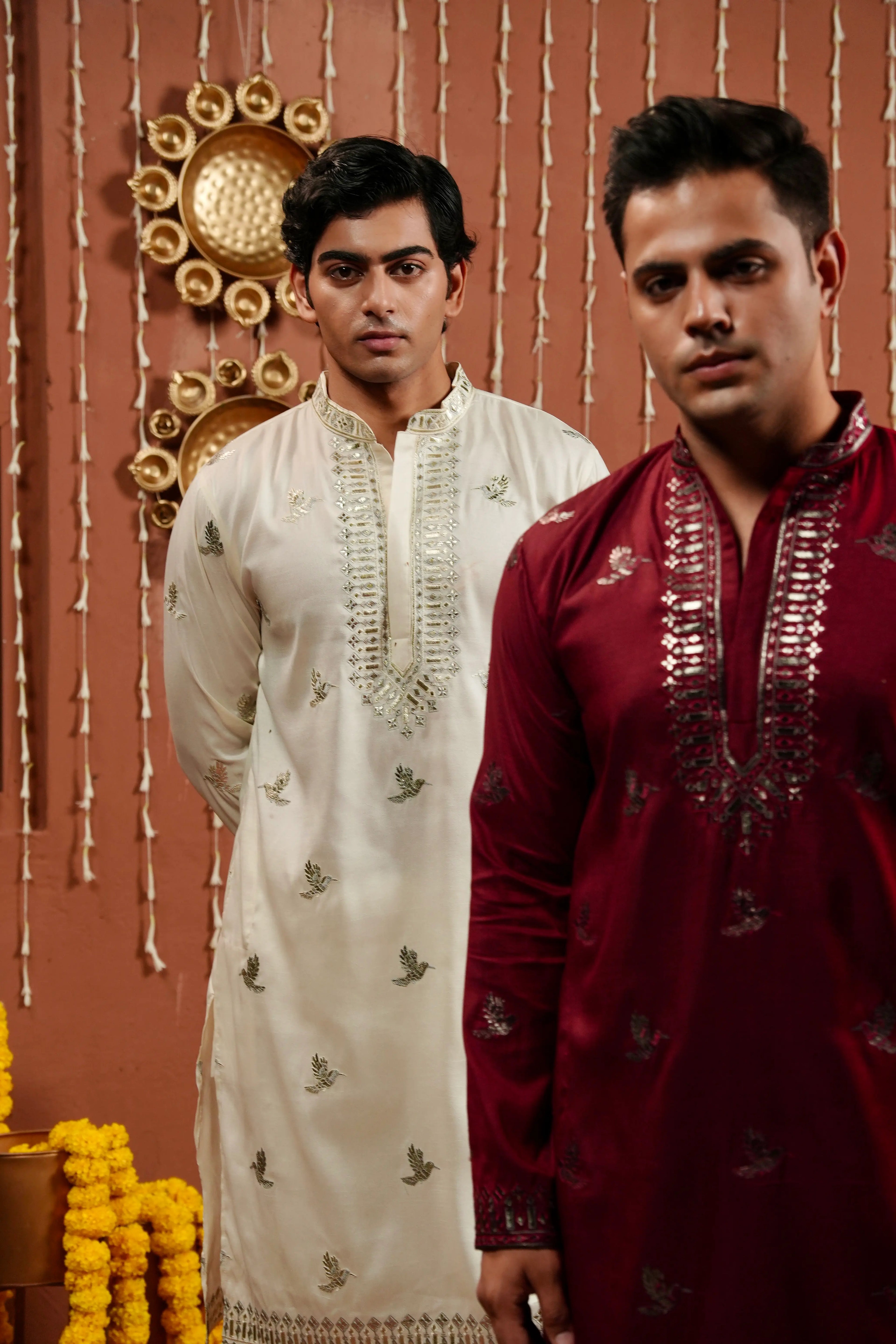 Two men wearing traditional Indian kurta sets, one in off-white with silver embroidery and the other in maroon with silver embroidery, standing against a decorated warm brown wall.