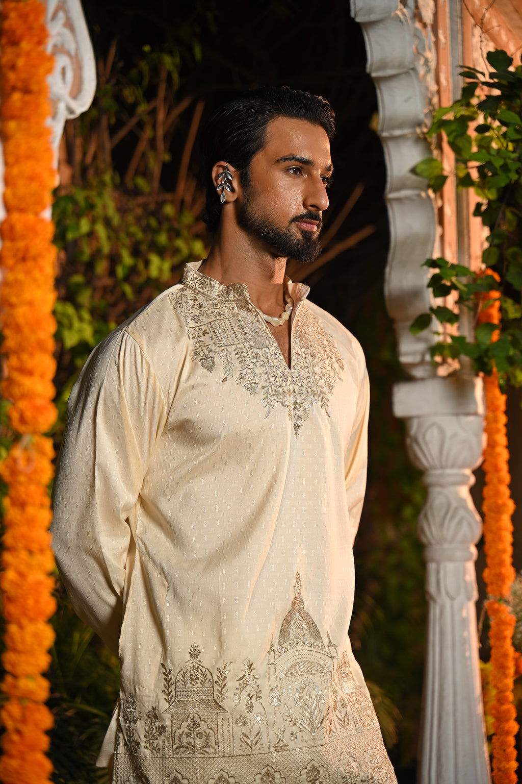 A man wearing a cream-colored embroidered kurta stands in an outdoor setting decorated with orange marigold garlands and white architectural pillars.